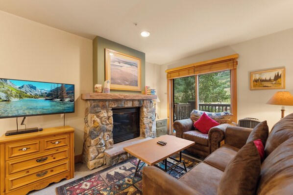 A cozy living room with a stone fireplace, a television, a comfortable couch, and a view of the outdoors through large windows. - Cozy Condo in River Run Village, King Bed, Walk to Gondola, Shops and Dining (Keystone)