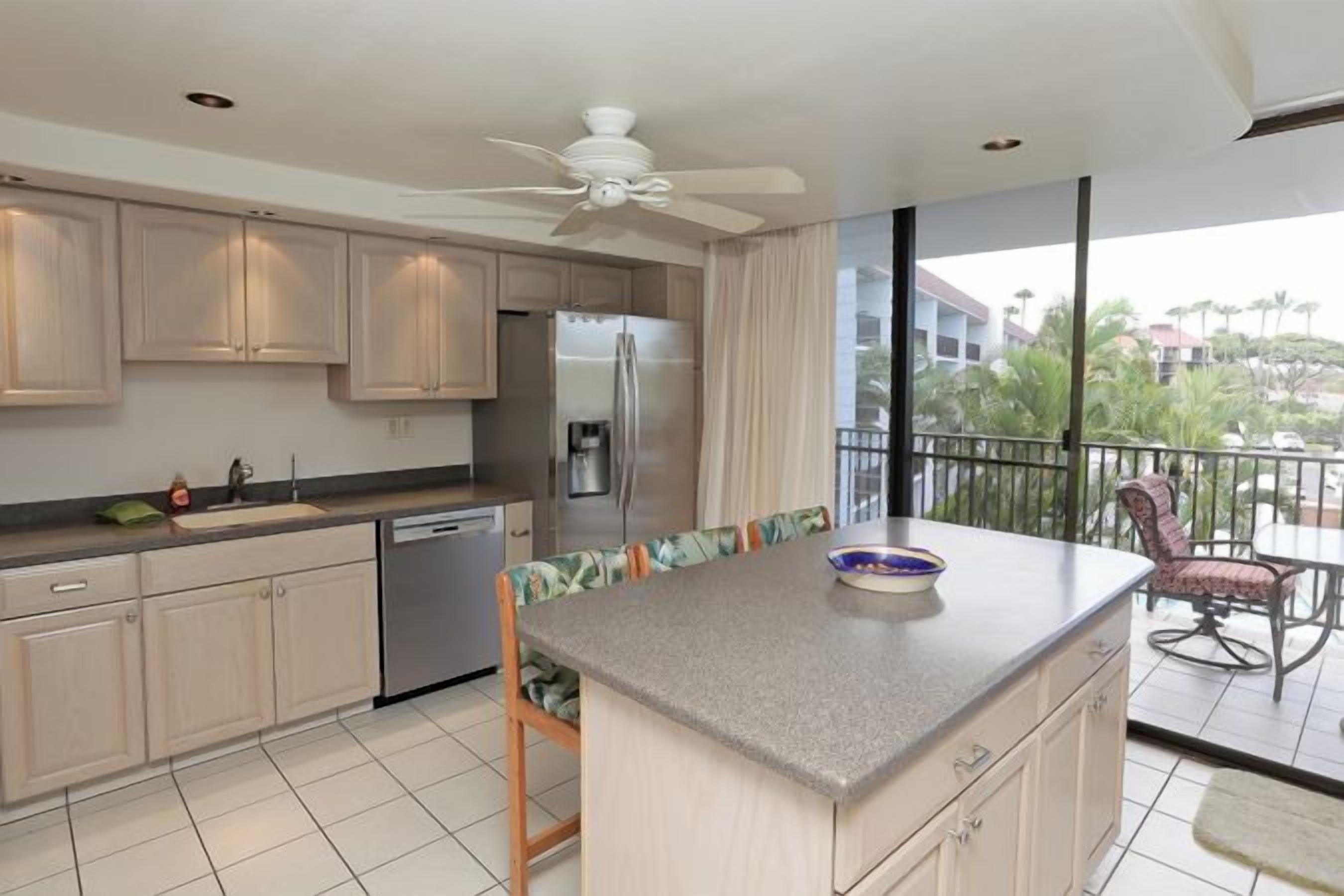 A modern kitchen with a central island, a refrigerator, and a dishwasher. There is a balcony with a view of palm trees and buildings.