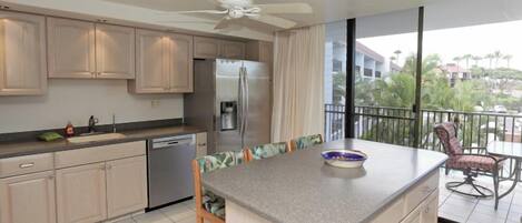 A modern kitchen with a central island, a refrigerator, and a dishwasher. There is a balcony with a view of palm trees and buildings.