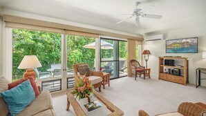A spacious living room with a large window, a ceiling fan, a television, and a bookshelf. - Manualoha 606 AC Throughout By Suite Paradise (Koloa)