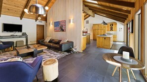 Ein großzügiger Wohnbereich mit einer Couch, einem Couchtisch und einem Esstisch mit Kerze. Es gibt eine Küchenzeile mit Holzschränken und einem an der Wand befestigten Fernseher.