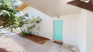A white brick building with a teal door, a small tree, and a concrete pathway.