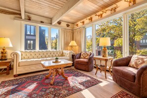 A spacious living room with a beige sofa, two armchairs, a coffee table, and a large window with a view of trees and buildings.