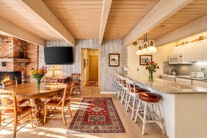 A kitchen with a wooden ceiling, a brick fireplace, a dining area with a table and chairs, and a bar with stools.
