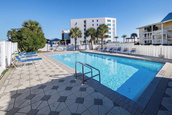 A swimming pool with lounge chairs, a white fence, and a building with multiple floors in the background.