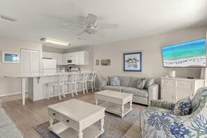 A modern kitchen with white cabinets, a dining area with white chairs, a gray sofa, and a flat-screen TV mounted on the wall.