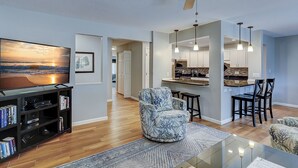 A modern living room with a flat-screen TV, a bookshelf, a patterned armchair, and a dining area with chairs and a table. - 105 Beachwalk – Pet-Friendly Comfort Just Steps from the Beach (Hilton Head Island)