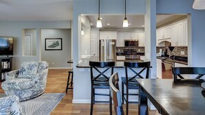 A modern kitchen with stainless steel appliances, a dining area with a black table and chairs, and a living room with a patterned sofa and a television. - 105 Beachwalk – Pet-Friendly Comfort Just Steps from the Beach (Hilton Head Island)