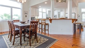A spacious dining area with a wooden table and chairs, a chandelier, and a view of the kitchen in the background. - Baywatch | Steps to the Sand, Game Room, Bay View (Tybee Island)