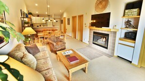 A living room with a fireplace, a television mounted above it, and a dining area with wooden furniture. - On River Trail! 2 Hot Tubs. Walk to Festivals, Dining, Shops, Activities (Keystone)