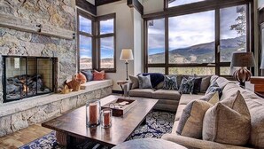 Un salón amplio con chimenea de piedra, ventanas grandes con vistas a la montaña, un conjunto de sofá acogedor y una mesa de centro de madera.
