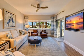 A modern living room with a sofa, armchairs, a coffee table, a ceiling fan, a TV, and a large window with a view of palm trees. - 433 Captains Walk- Completely Renovated - Oceanfront Villa in Palmetto Dunes! (Hilton Head Island)