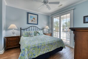 Une chambre à coucher comprenant un lit, deux tables de chevet, un ventilateur de plafond, une grande fenêtre et une porte donnant sur un balcon.