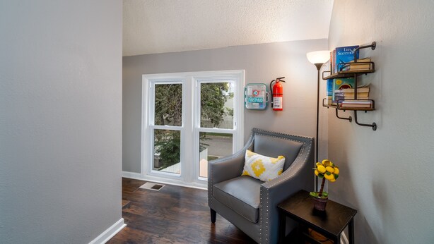 A room with a gray armchair, a small table with a plant, a wall-mounted bookshelf, and a fire extinguisher.