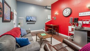 A living room with a grey sofa, a coffee table, a red accent wall, a kitchen area with a refrigerator, and a dining table with stools.