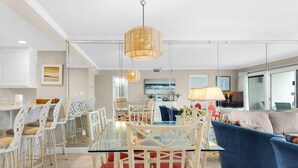 A modern living room with a glass dining table, white chairs, a blue sofa, and a large window.