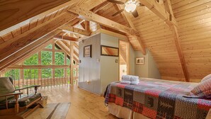 Een gezellig slaapkamer op zolder met een houten plafond, een groot bed met een kleurrijk sprei, een houten stoel en uitzicht op groen buiten.