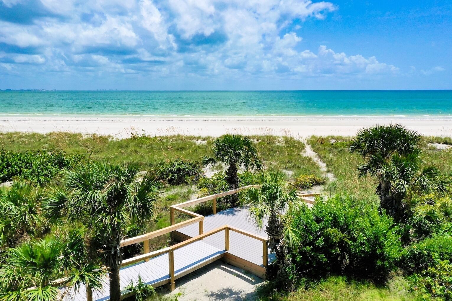 A beach with a wooden boardwalk leading to a sandy area, surrounded by greenery and palm trees.
