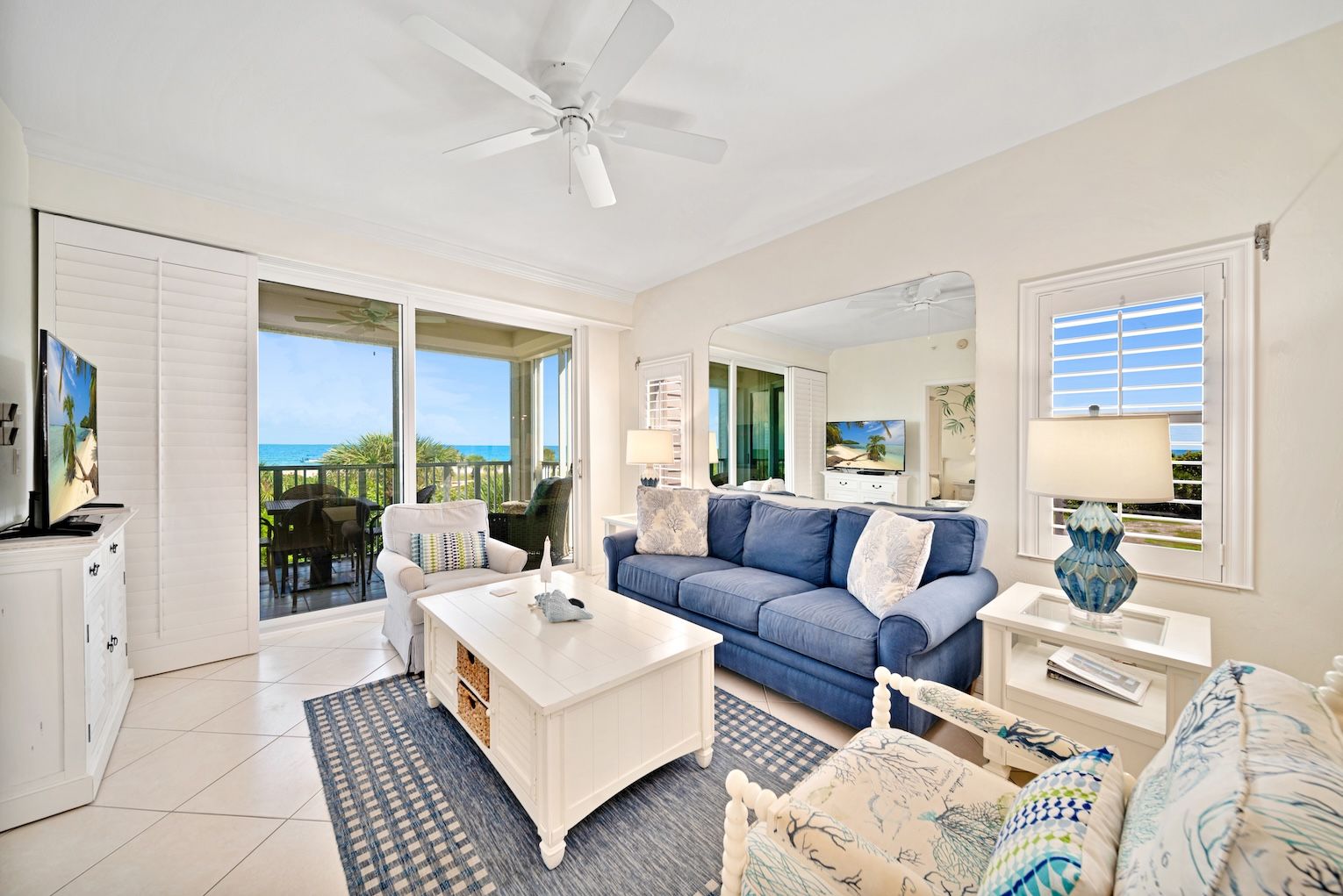 A spacious living room with a large sofa, a coffee table, and a TV. There is a balcony with a view of the ocean.