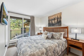 A bedroom with a large bed, a painting, a TV, and a balcony with a view of trees.
