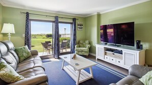A living room with a sofa, armchair, and coffee table, a TV, and a view of a green landscape through large windows.