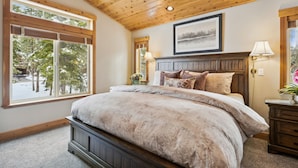 A bedroom with a wooden ceiling, a large window with blinds, a bed with a beige comforter, a bedside table with a lamp, and a framed picture on the wall.