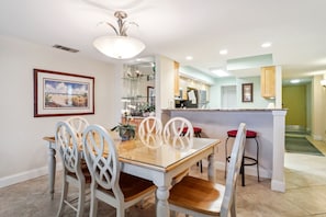 Uma sala de jantar com mesa de madeira e cadeiras brancas, uma cozinha com bancada e um corredor que dá para outro quarto.