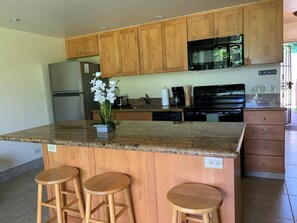 A kitchen with wooden cabinets, a granite countertop, a refrigerator, a stove, and a sink. There are three wooden stools at the island.