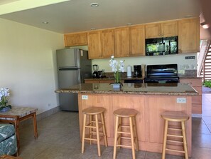 A modern kitchen with wooden cabinets, a stainless steel refrigerator, a microwave, a stove, and a blender. There are two wooden stools at the island.