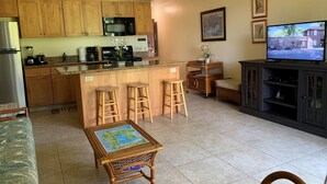 A compact kitchen with wooden cabinets, a microwave, and a coffee maker. There is a small dining area with a table and two stools. A television is mounted on the wall, and a wicker chair is visible. - Waiohuli Beach Hale #D-120 Steps to Beach Oceanfront Complex with Great Rates (Kihei)