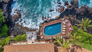 Vista aérea de un resort con piscina rodeada de sillones de playa y sombrillas, con vistas a un litoral rocoso y el océano.