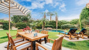 A wooden outdoor dining set with striped cushions, a striped umbrella, and a pool in the background. - Firefly by AvantStay | Spacious Backyard w/ Pool& Hot Tub! (La Quinta)