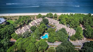 Vista aerea di un resort costiero con diversi edifici in stile bungalow, una piscina centrale e una spiaggia sullo sfondo.