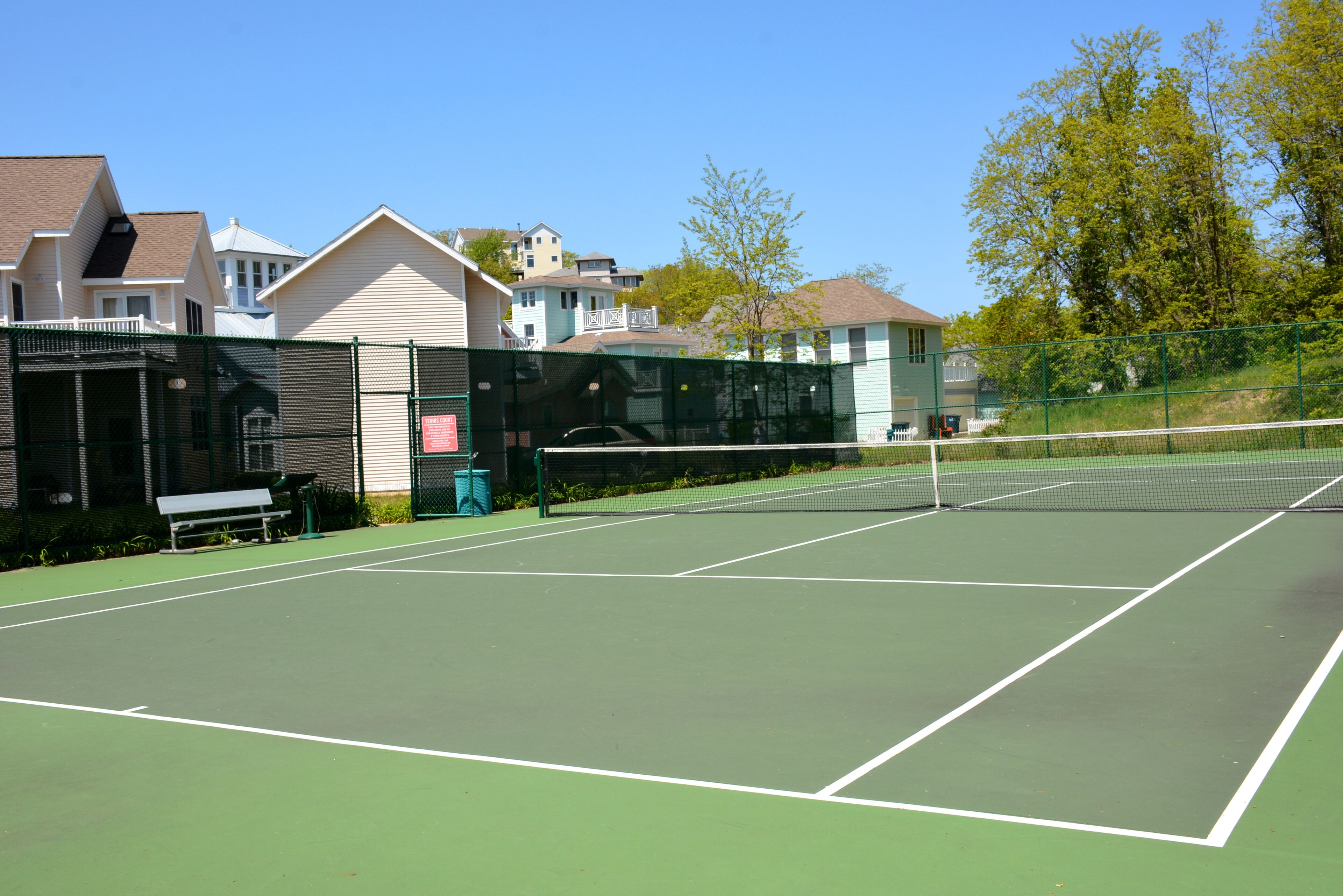 A tennis court with a net, surrounded by a fence, and houses in the background.