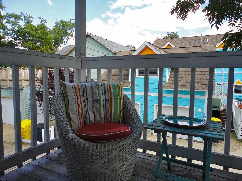 Une chaise en osier avec un coussin rayé, posée sur une terrasse en bois, une petite table avec une assiette, et en arrière-plan, la vue de bâtiments aux couleurs vives.