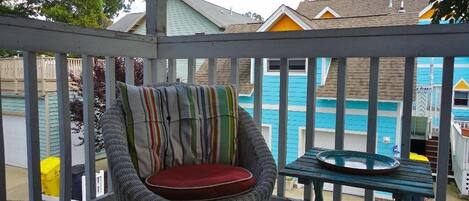 Une chaise en osier avec un coussin rayé, posée sur une terrasse en bois, une petite table avec une assiette, et en arrière-plan, la vue de bâtiments aux couleurs vives.
