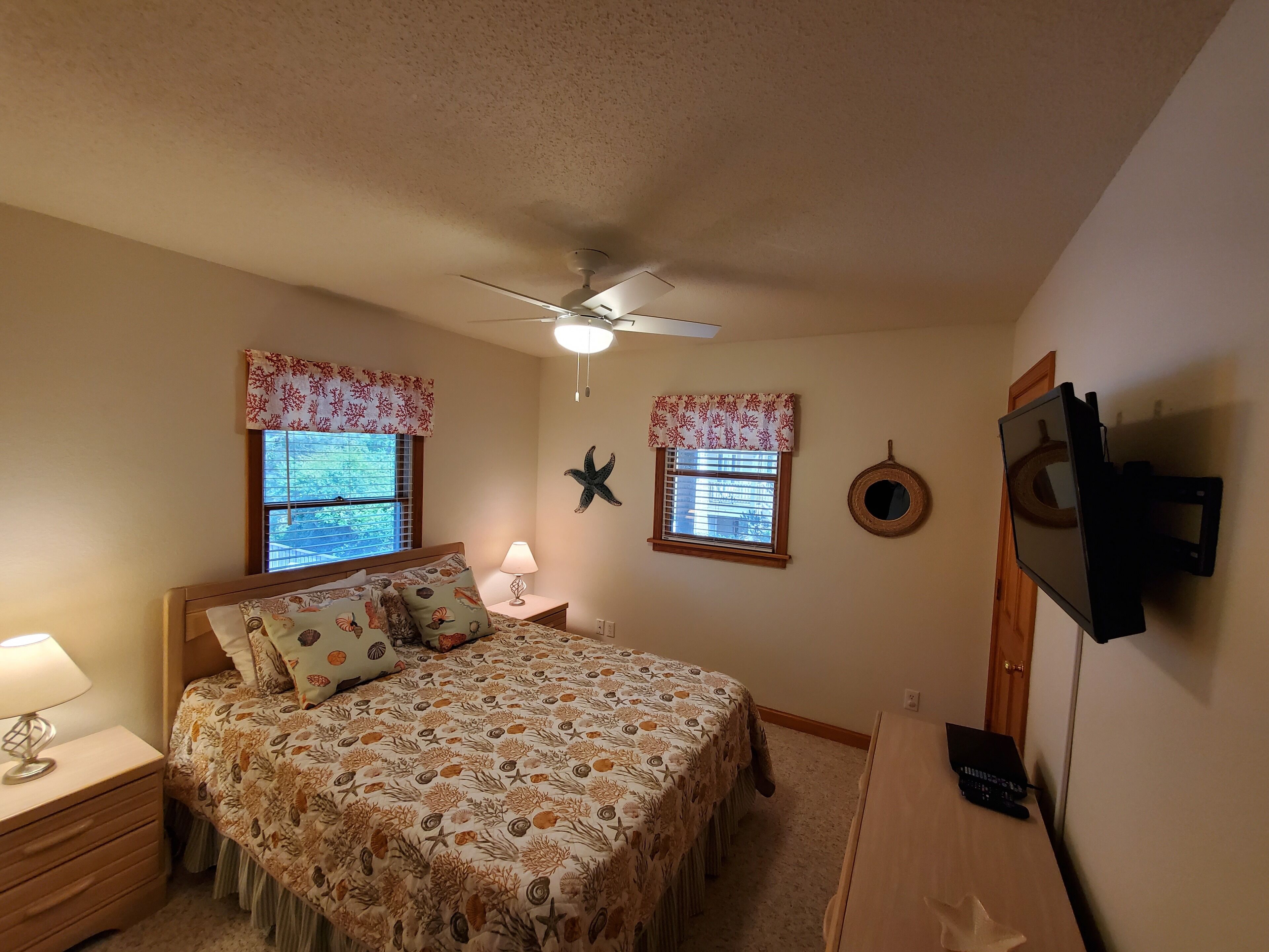 A bedroom with a bed, two windows with floral valances, a ceiling fan, a wall-mounted TV, and a starfish decoration.