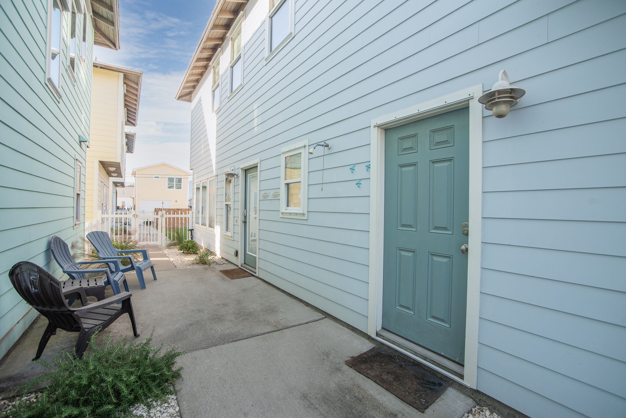 A narrow residential street with light blue houses, a green door, and outdoor seating.