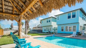 A backyard with a swimming pool, a thatched roof structure, and a two-story house. - Pura Vida Cottage! Community Pool, Downtown PA! (Port Aransas)