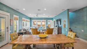 A spacious dining area with a wooden table and yellow chairs, a kitchen in the background, and a view of the outdoors through large windows. - Pura Vida Cottage! Community Pool, Downtown PA! (Port Aransas)