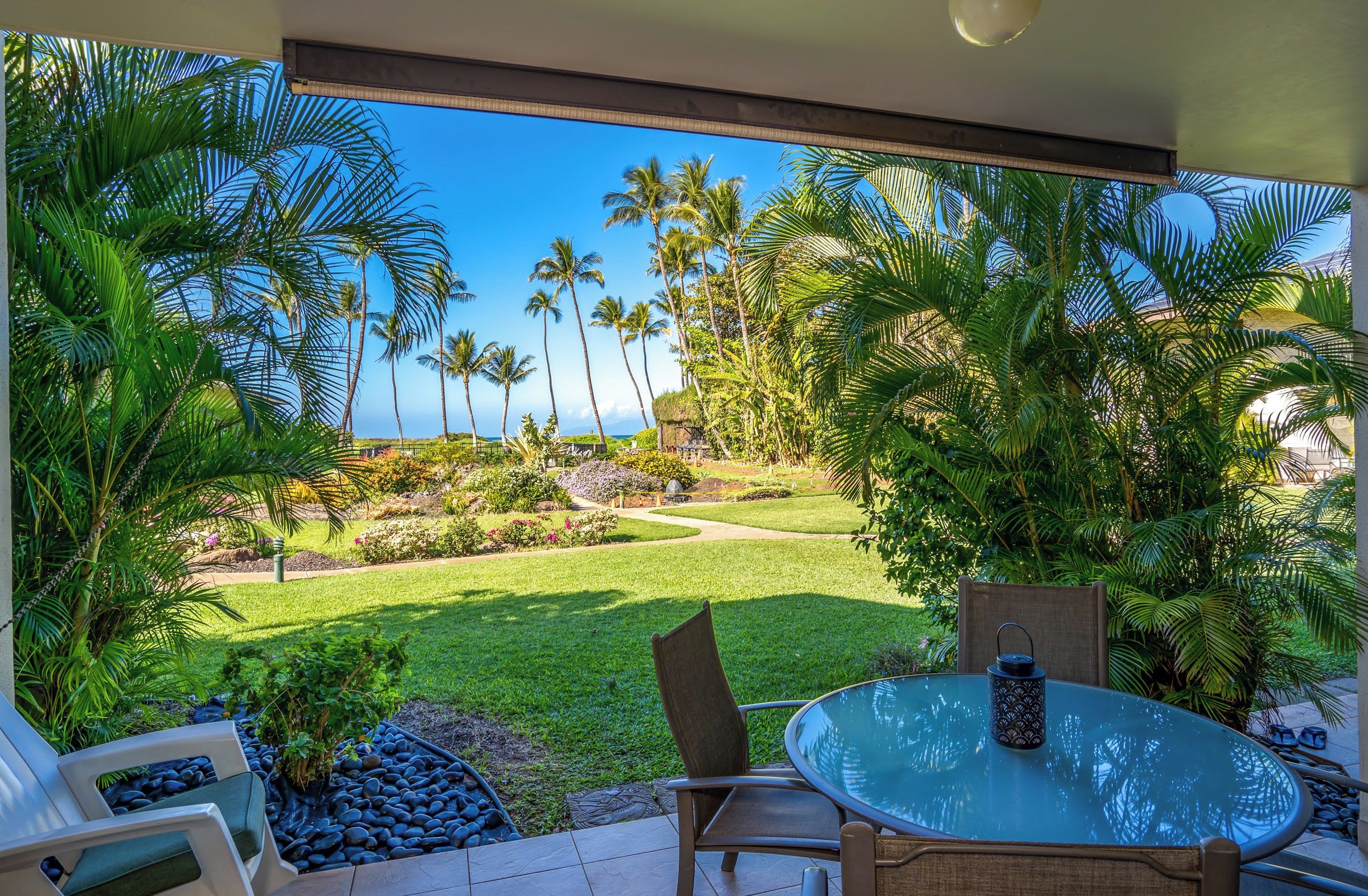 A patio with a glass table and chairs, overlooking a well-maintained garden with palm trees and a view of the ocean.