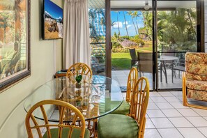 A dining area with a glass table and wicker chairs, a view of a tropical garden through a sliding glass door, and a framed picture on the wall.