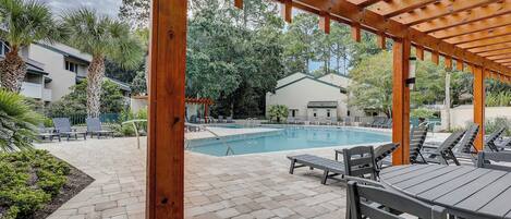 A covered outdoor pool area with lounge chairs and a table, surrounded by greenery and buildings.