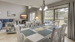 Een eethoek met een tafel voor vier personen, omringd door witte stoelen, een kroonluchter en een uitzicht op de omgeving door grote ramen.