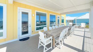 A covered outdoor dining area with a wooden table and white chairs, large windows, and a view of the ocean.