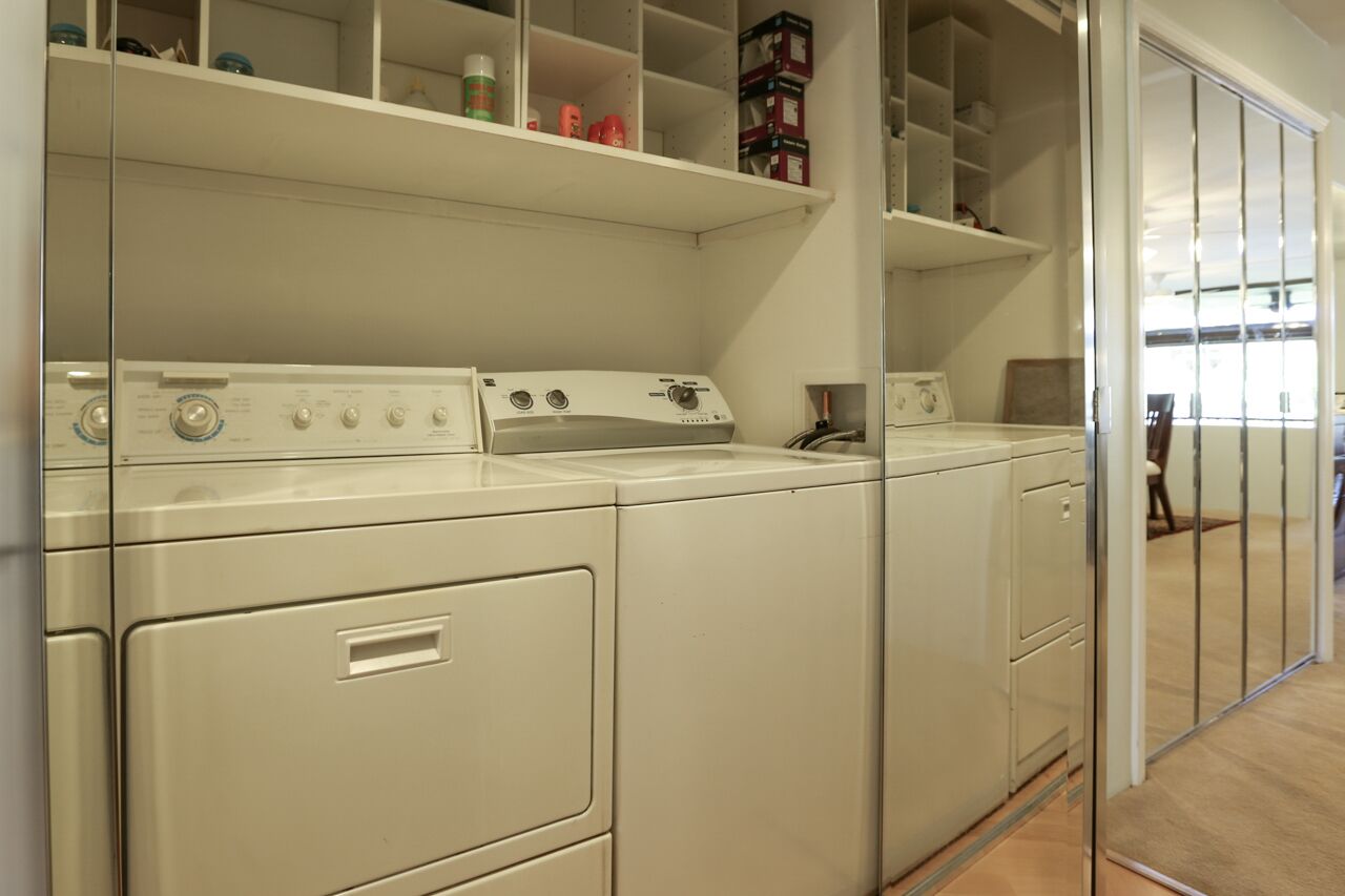 Een wasruimte met een voorlader wasmachine en droger, boven liggen planken met opbergdozen, en een schuifdeur die leidt naar een andere ruimte.