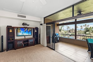 A living room with a large flat-screen TV, a wooden entertainment unit, and a balcony with a view of palm trees.