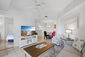 A modern living room with a flat-screen TV, a coffee table, a sofa, a dining area with a table and chairs, and a ceiling fan.