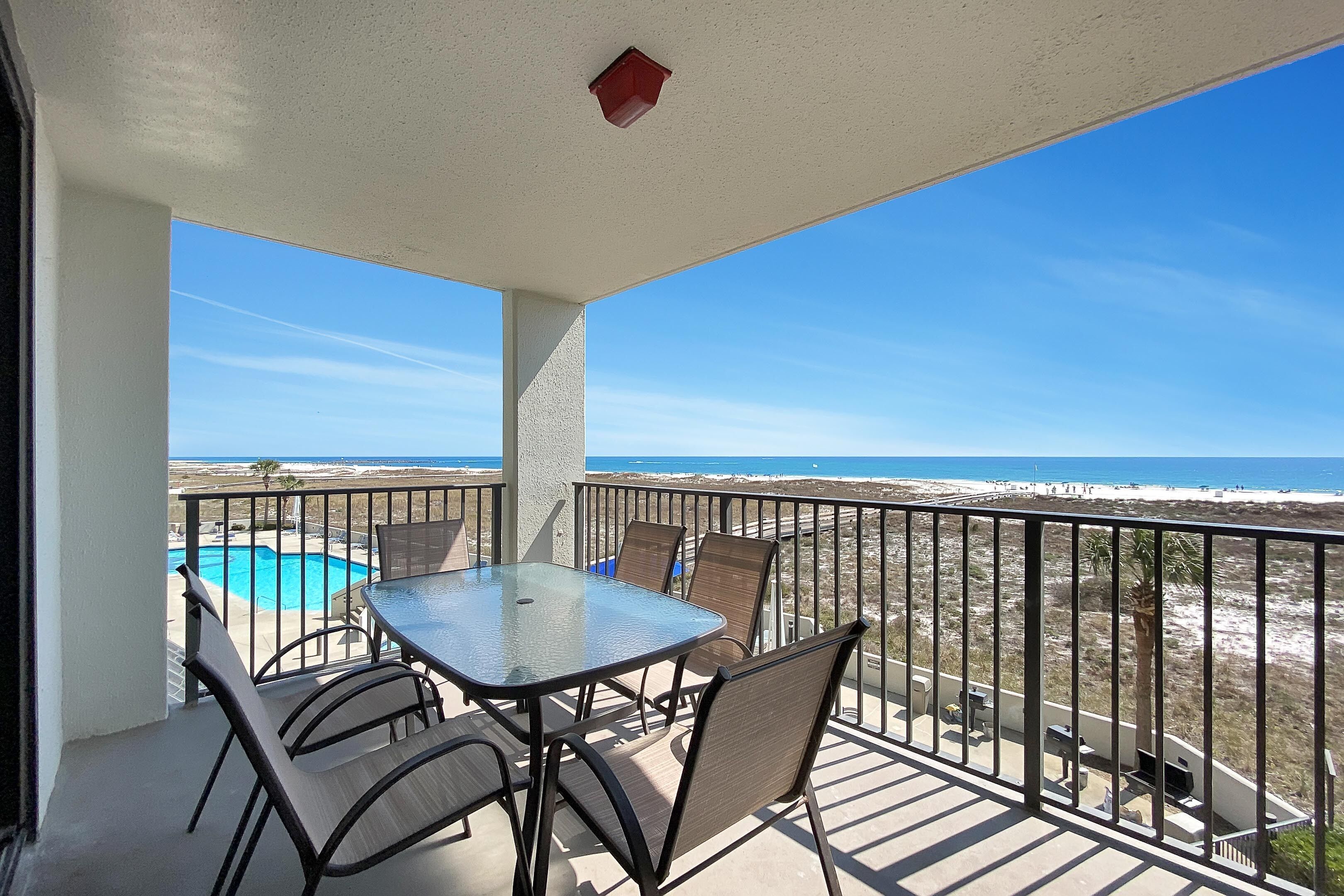 A balcony with a glass table and chairs, overlooking a beach and ocean.