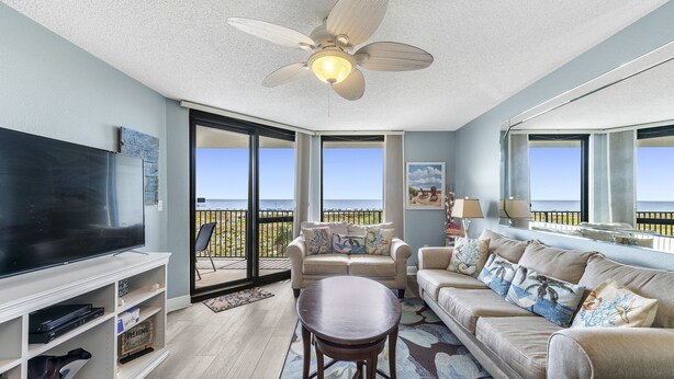 A spacious living room with a large flat-screen TV, a ceiling fan, and a view of the ocean through large sliding glass doors. - Phoenix VI 204 | Beachfront | Resort Amenities | My Beach Getaways (Orange Beach)